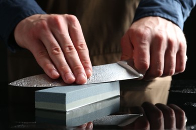 Photo of Man sharpening knife with sharpener at mirror table, closeup