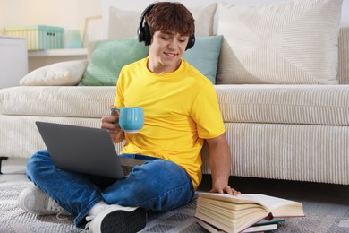 Photo of Teenage student with cup of drink and laptop studying on floor at home