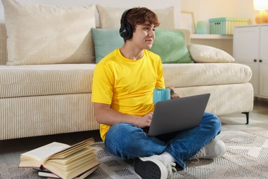 Photo of Teenage student with cup of drink using laptop while studying on floor at home