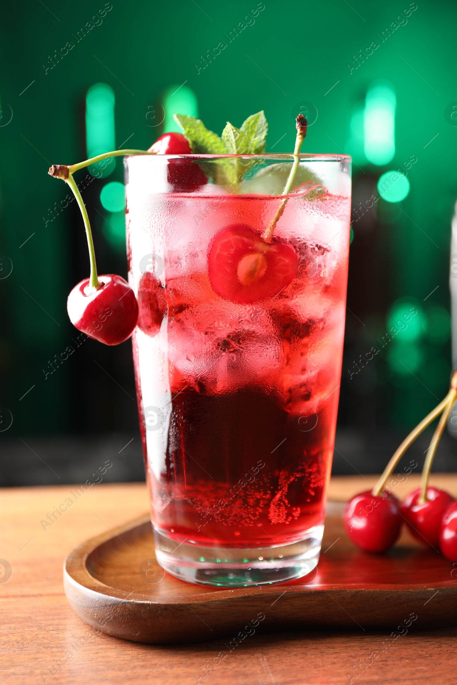 Tasty cherry soda water with ice cubes, berries and mint in glass on table against blurred green background, closeup Photo of Tasty cherry soda water with ice cubes, berries and mint in glass on table against blurred green background, closeup