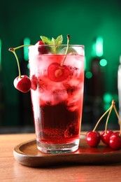 Photo of Tasty cherry soda water with ice cubes, berries and mint in glass on table against blurred green background, closeup