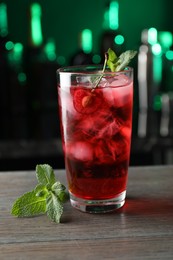 Photo of Tasty cherry soda water with ice cubes, berry and mint in glass on wooden table against blurred green background, closeup