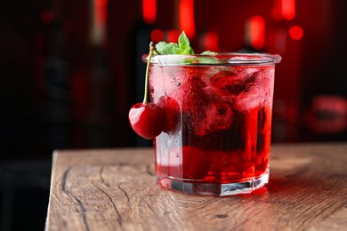 Photo of Tasty cherry soda water with ice cubes, berries and mint in glass on wooden table against blurred background, closeup. Space for text