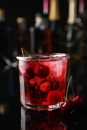 Photo of Tasty cherry soda water with ice cubes and berries in glass on mirror table against blurred background, closeup