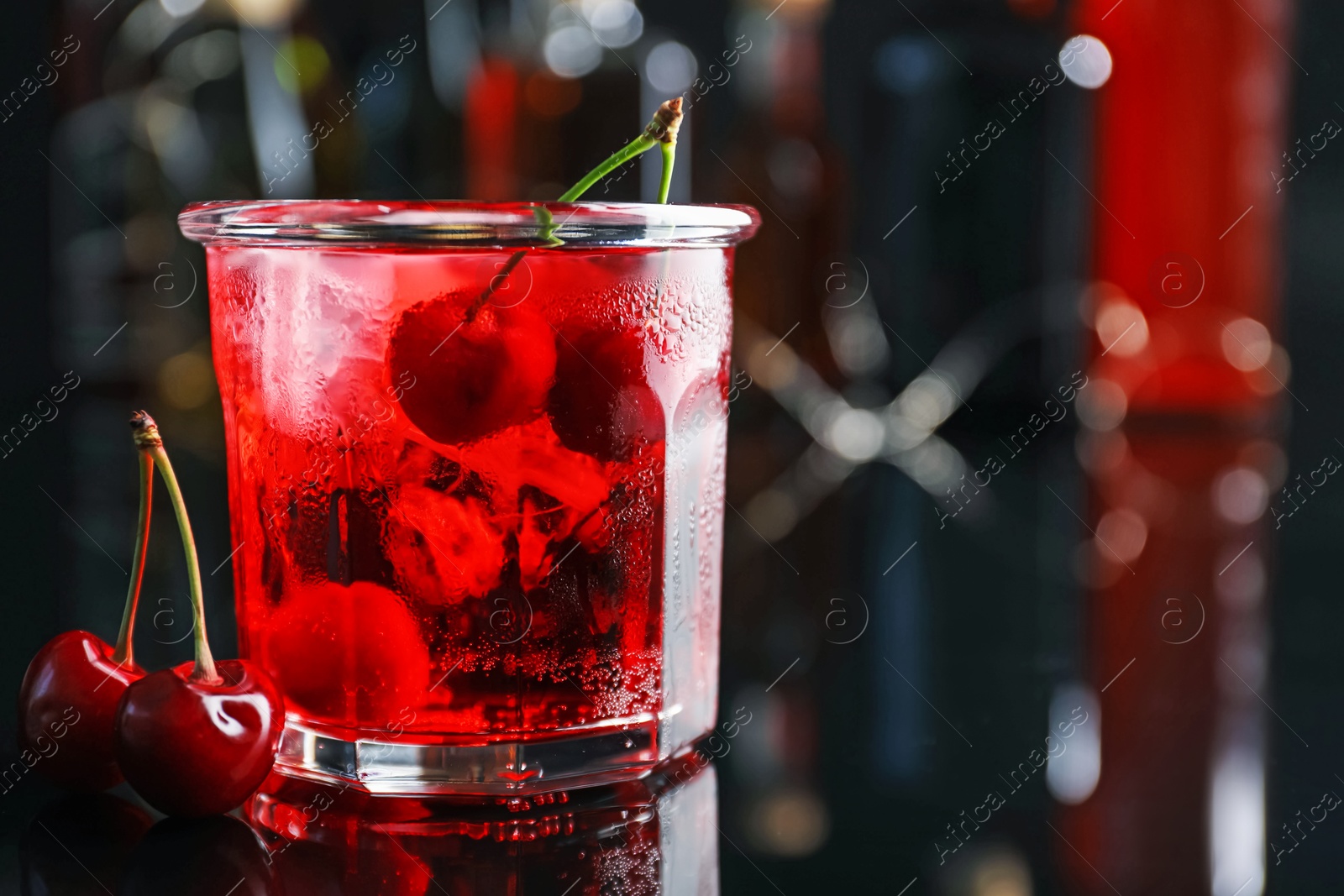 Photo of Tasty cherry soda water with ice cubes and berries in glass on mirror table against blurred background, closeup