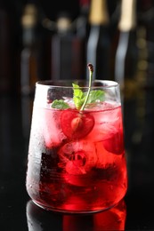 Photo of Tasty cherry soda water with ice cubes, berry and mint in glass on mirror table against blurred background, closeup