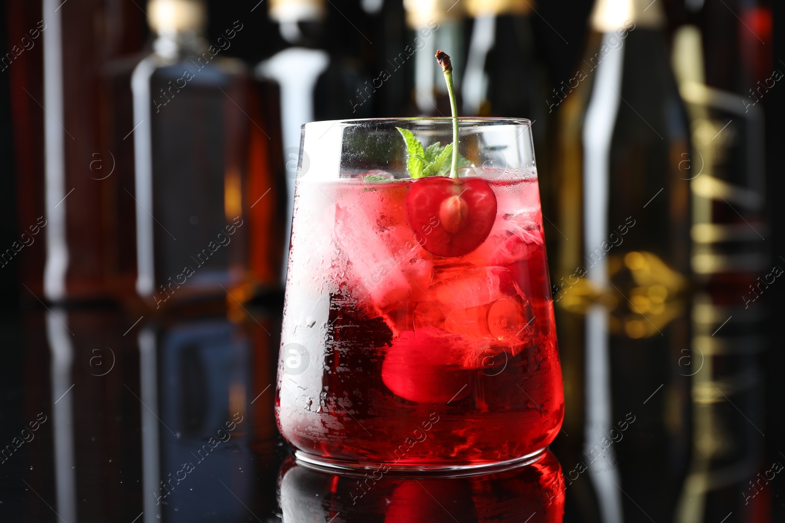 Tasty cherry soda water with ice cubes, berry and mint in glass on mirror table against blurred background, closeup Photo of Tasty cherry soda water with ice cubes, berry and mint in glass on mirror table against blurred background, closeup