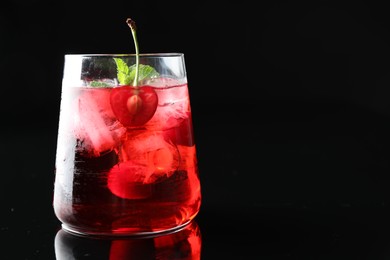 Photo of Tasty cherry soda water with ice cubes, berry and mint in glass on mirror table against black background, closeup. Space for text