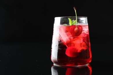 Photo of Tasty cherry soda water with ice cubes, berry and mint in glass on mirror table against black background, closeup. Space for text
