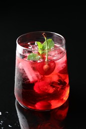 Photo of Tasty cherry soda water with ice cubes, berry and mint in glass on mirror table against black background, closeup