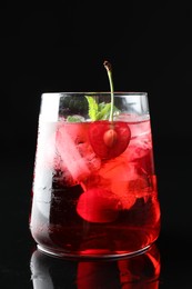 Photo of Tasty cherry soda water with ice cubes, berry and mint in glass on mirror table against black background, closeup