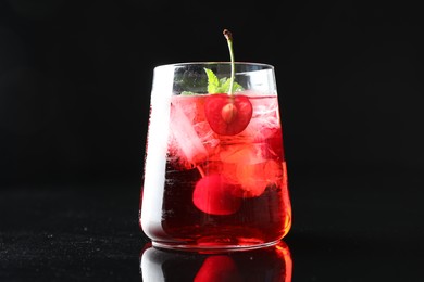 Photo of Tasty cherry soda water with ice cubes, berry and mint in glass on mirror table against black background, closeup