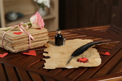 Photo of Stack of love letters, parchment sheet, ink, quill, rose flower and red hearts on wooden table indoors, closeup