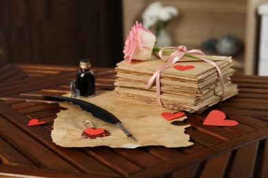 Photo of Stack of love letters, parchment sheet, ink, quill, rose flower and red hearts on wooden table indoors, closeup