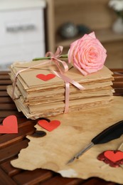 Photo of Stack of love letters, parchment sheet, quill, rose flower and red hearts on wooden table indoors, closeup