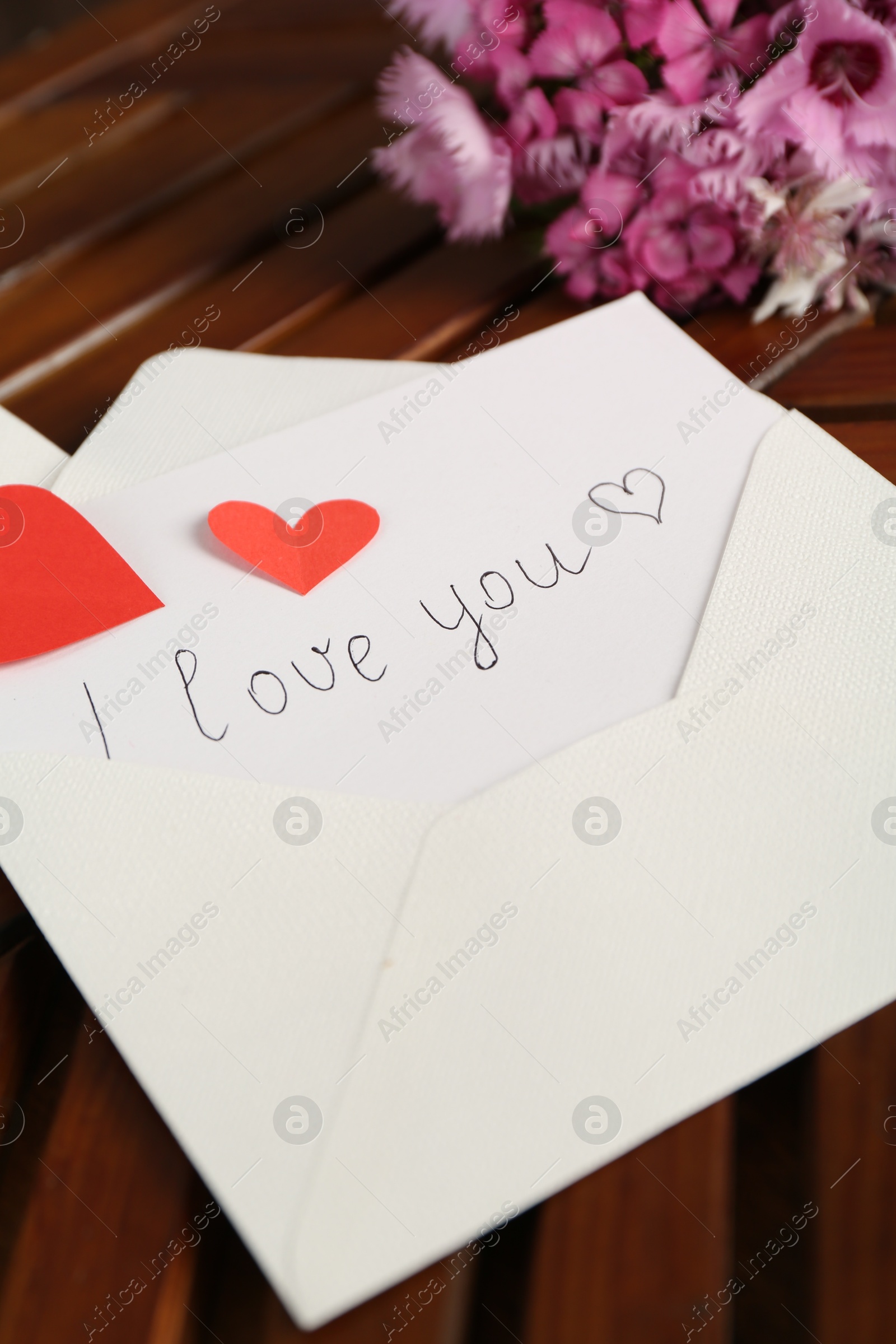 Photo of Card with phrase I Love You, envelope, red hearts and flowers on wooden table indoors, closeup