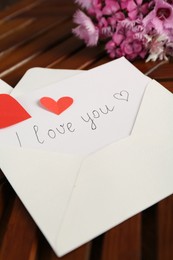 Photo of Card with phrase I Love You, envelope, red hearts and flowers on wooden table indoors, closeup