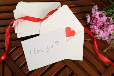 Photo of Card with phrase I Love You, letters, red heart, ribbon and flowers on wooden table, flat lay