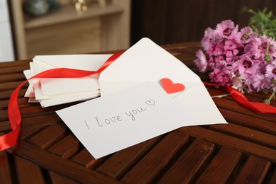 Photo of Card with phrase I Love You, letters, red heart, ribbon and flowers on wooden table indoors, closeup