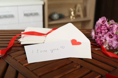 Photo of Card with phrase I Love You, letters, red heart, ribbon and flowers on wooden table indoors, closeup