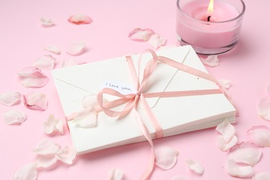Photo of Stack of letters with phrase I Love You, burning candle and petals on pink background, closeup