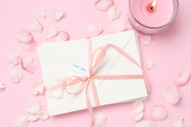Photo of Stack of letters with phrase I Love You, burning candle and petals on pink background, flat lay