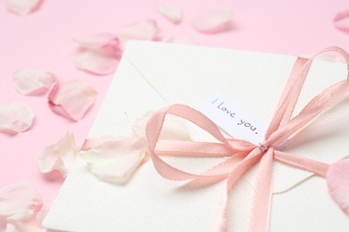 Photo of Envelope with phrase I Love You and petals on pink background, closeup