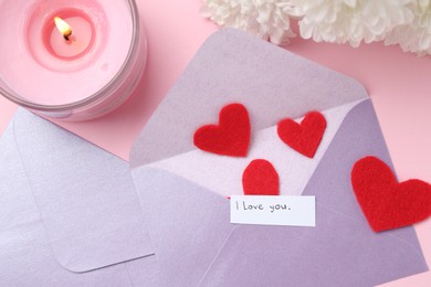 Photo of Envelopes with phrase I Love You, red hearts, burning candle and flowers on pink background, flat lay