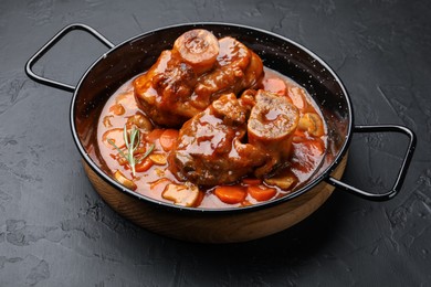 Photo of Tasty beef ossobuco with carrot, mushrooms and rosemary in frying pan on dark textured table, closeup
