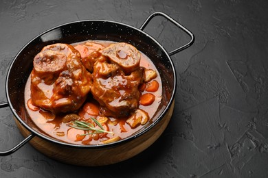 Photo of Tasty beef ossobuco with carrot, mushrooms and rosemary in frying pan on dark textured table, closeup