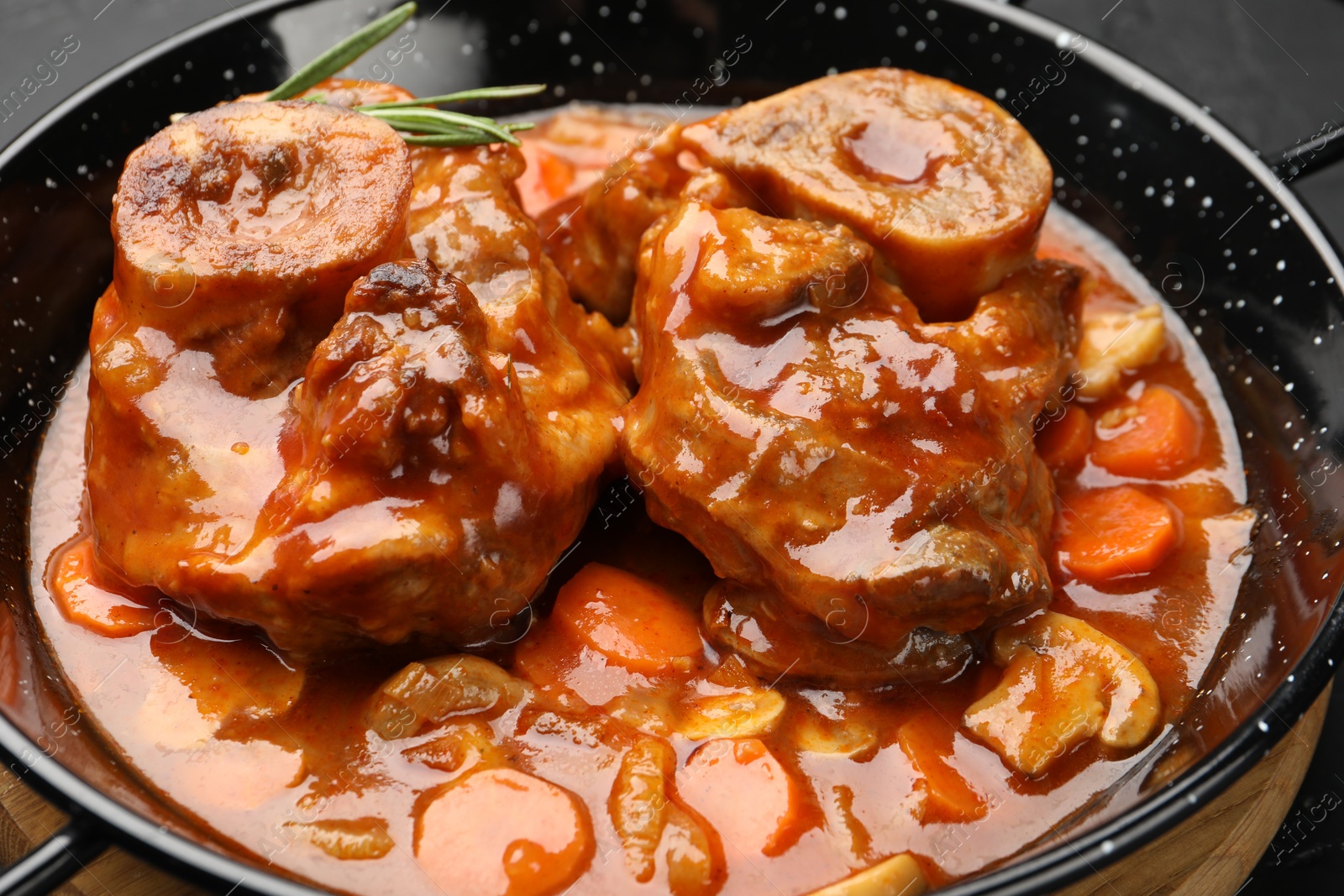 Tasty beef ossobuco with carrot, mushrooms and rosemary in frying pan on table, closeup Photo of Tasty beef ossobuco with carrot, mushrooms and rosemary in frying pan on table, closeup