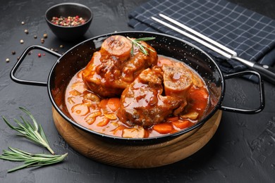Photo of Tasty beef ossobuco with carrot, mushrooms and rosemary in frying pan on dark textured table, closeup