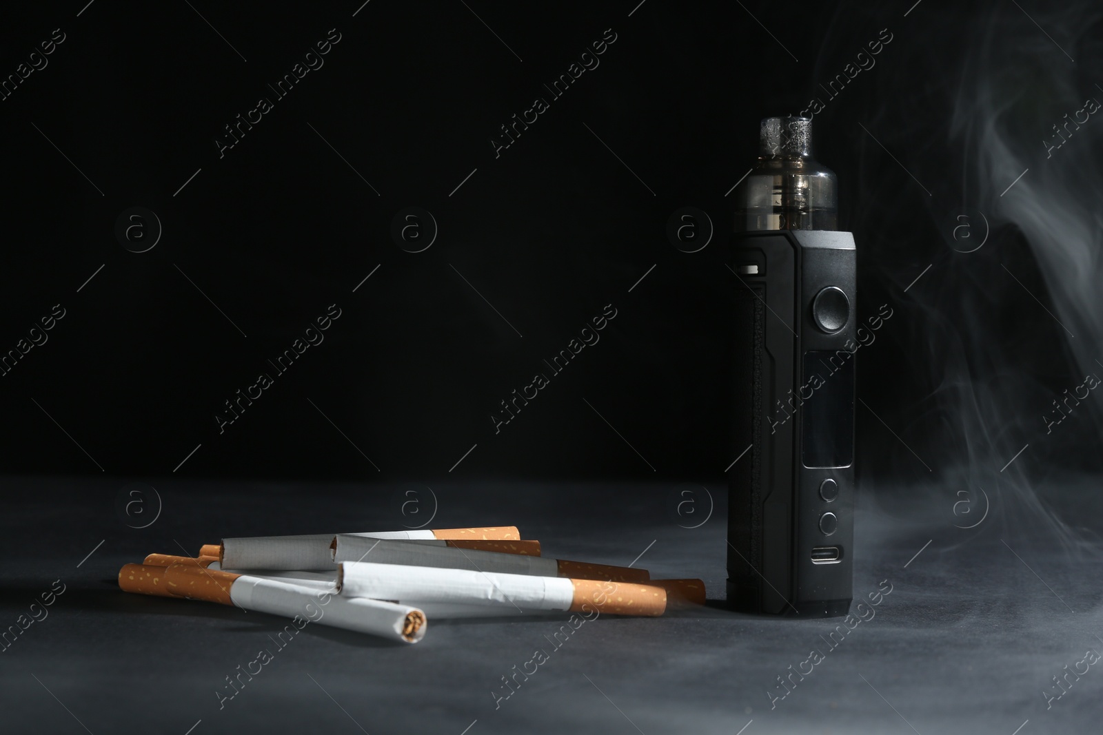 Electronic smoking device and cigarettes in smoke on dark table against black background, closeup Photo of Electronic smoking device and cigarettes in smoke on dark table against black background, closeup