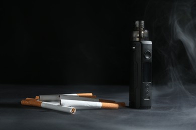 Photo of Electronic smoking device and cigarettes in smoke on dark table against black background, closeup