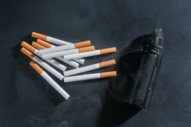 Photo of Electronic smoking device and cigarettes in smoke on dark table, flat lay