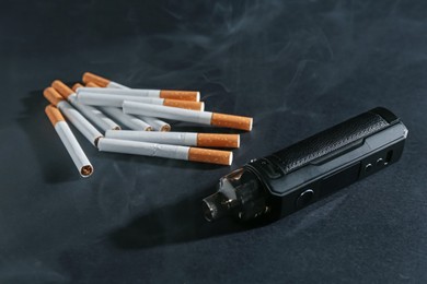 Photo of Electronic smoking device and cigarettes in smoke on dark table, closeup
