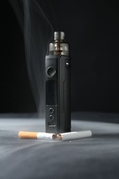 Photo of Electronic smoking device and broken cigarette in smoke on dark table against black background, closeup