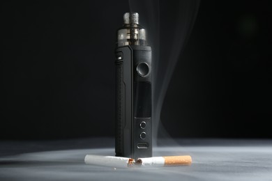 Photo of Electronic smoking device and broken cigarette in smoke on dark table against black background, closeup