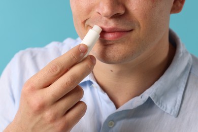 Photo of Young man applying lip balm onto his lips on light blue background, closeup