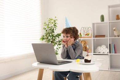 Photo of Birthday celebration. Little boy with cupcake having video call via laptop at table indoors