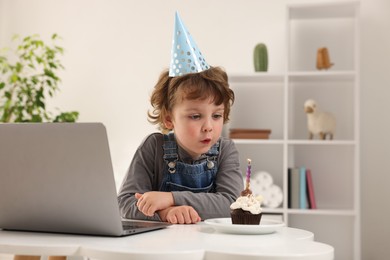 Photo of Birthday celebration. Little boy blowing candle on cupcake while having video call via laptop at table indoors