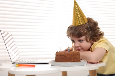 Photo of Birthday celebration. Little boy with cake having video call via laptop at table indoors. Space for text