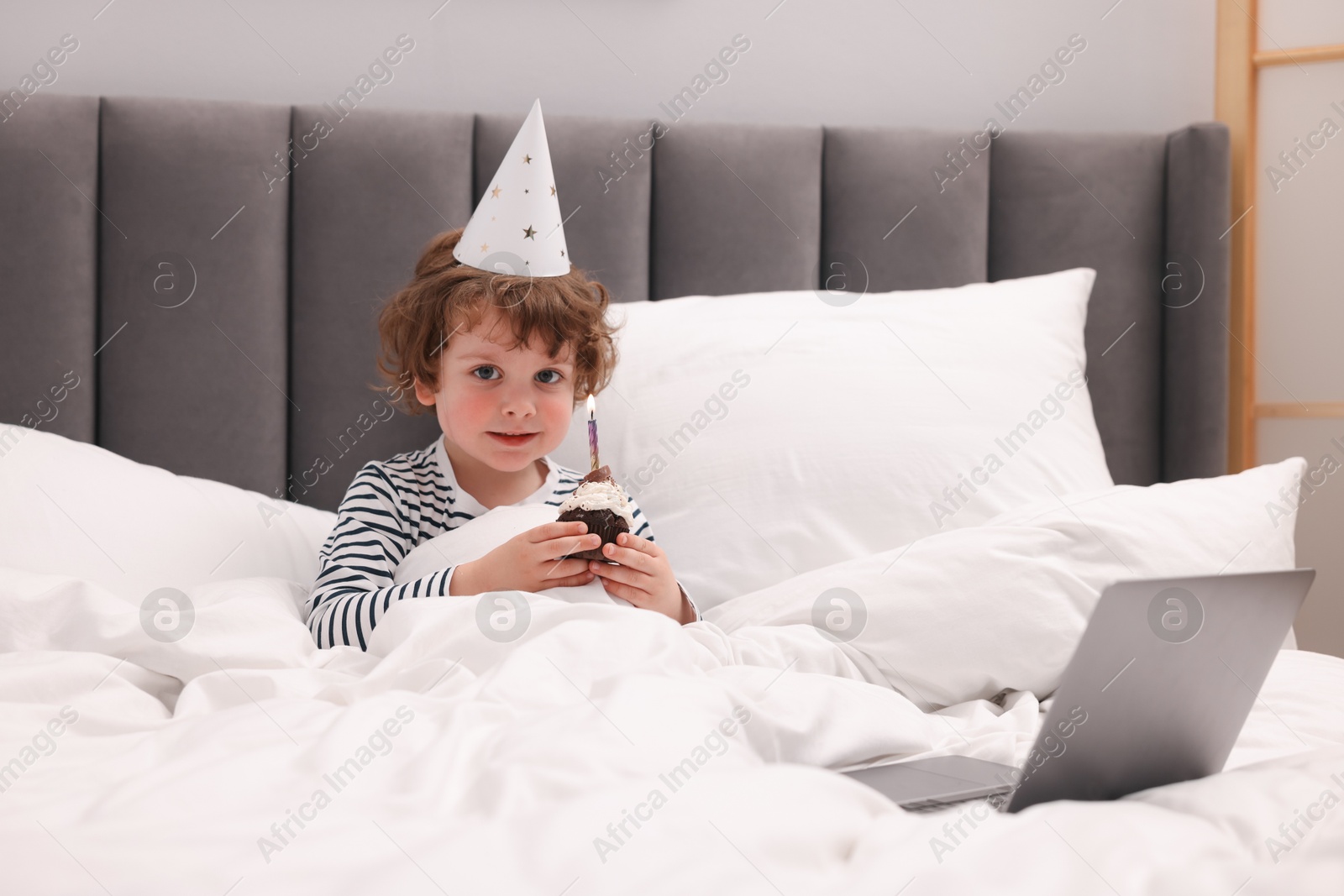 Birthday celebration. Little boy with cupcake having video call via laptop in bed at home Photo of Birthday celebration. Little boy with cupcake having video call via laptop in bed at home