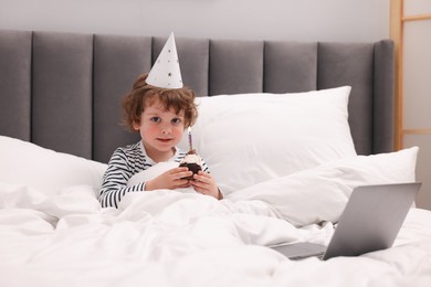 Photo of Birthday celebration. Little boy with cupcake having video call via laptop in bed at home
