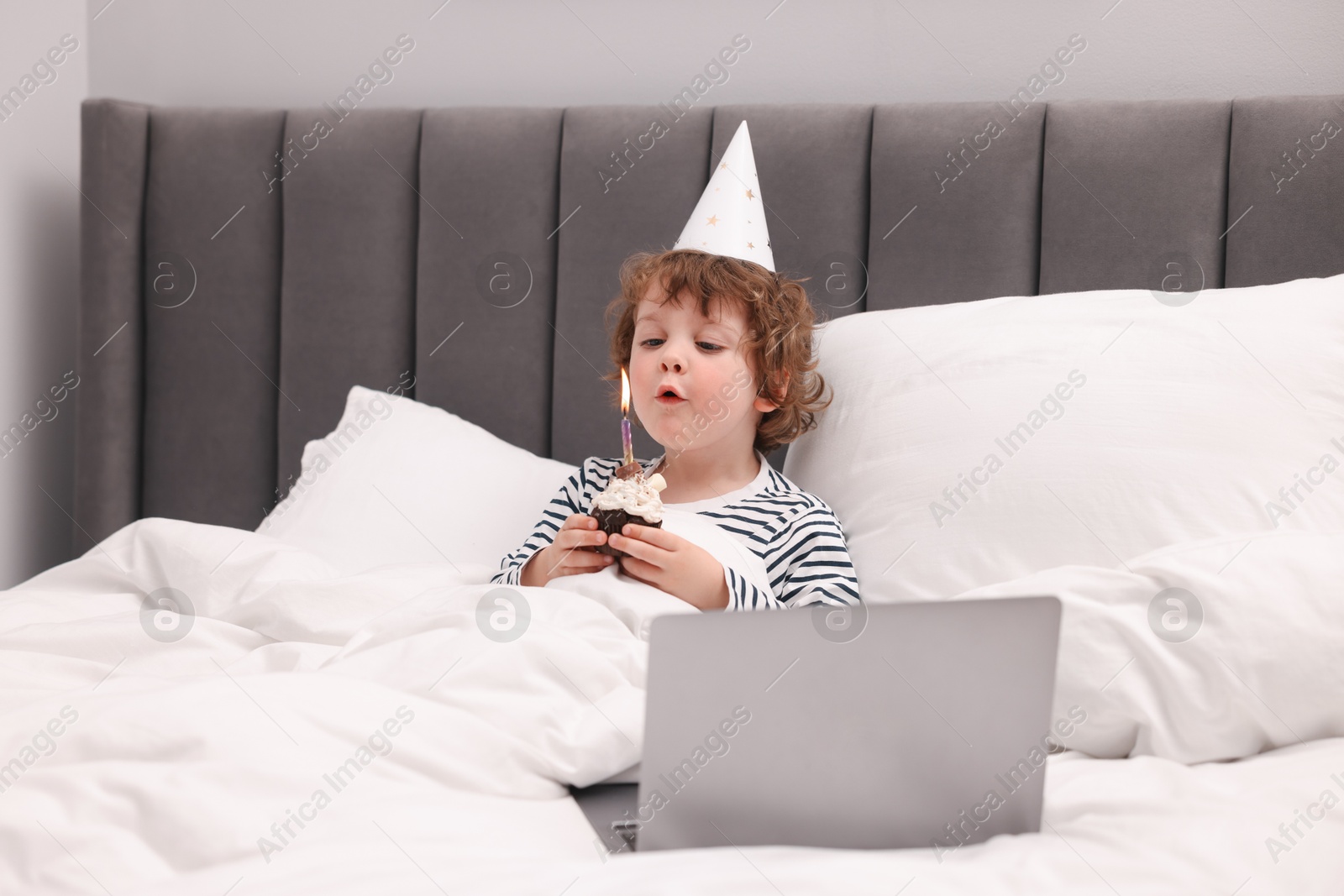 Birthday celebration. Little boy blowing candle on cupcake while having video call via laptop in bed at home Photo of Birthday celebration. Little boy blowing candle on cupcake while having video call via laptop in bed at home