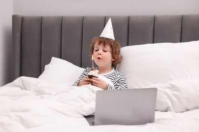 Photo of Birthday celebration. Little boy blowing candle on cupcake while having video call via laptop in bed at home