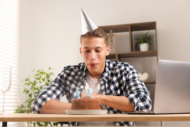 Photo of Birthday celebration. Man blowing candles on cake while having video call via laptop at desk indoors