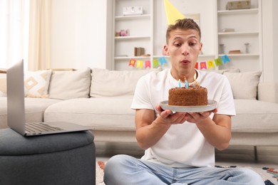 Photo of Birthday celebration. Man blowing candles on cake while having video call via laptop at home