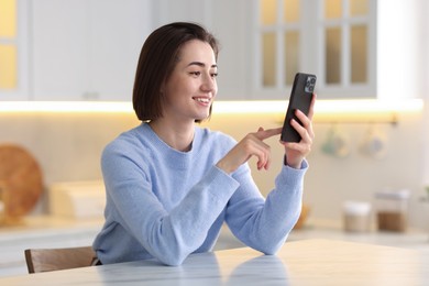 Photo of Woman using modern smartphone at table in kitchen