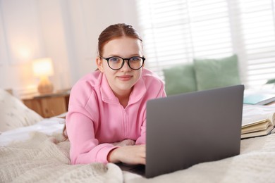 Photo of Teenage student in glasses using laptop while studying on bed at home